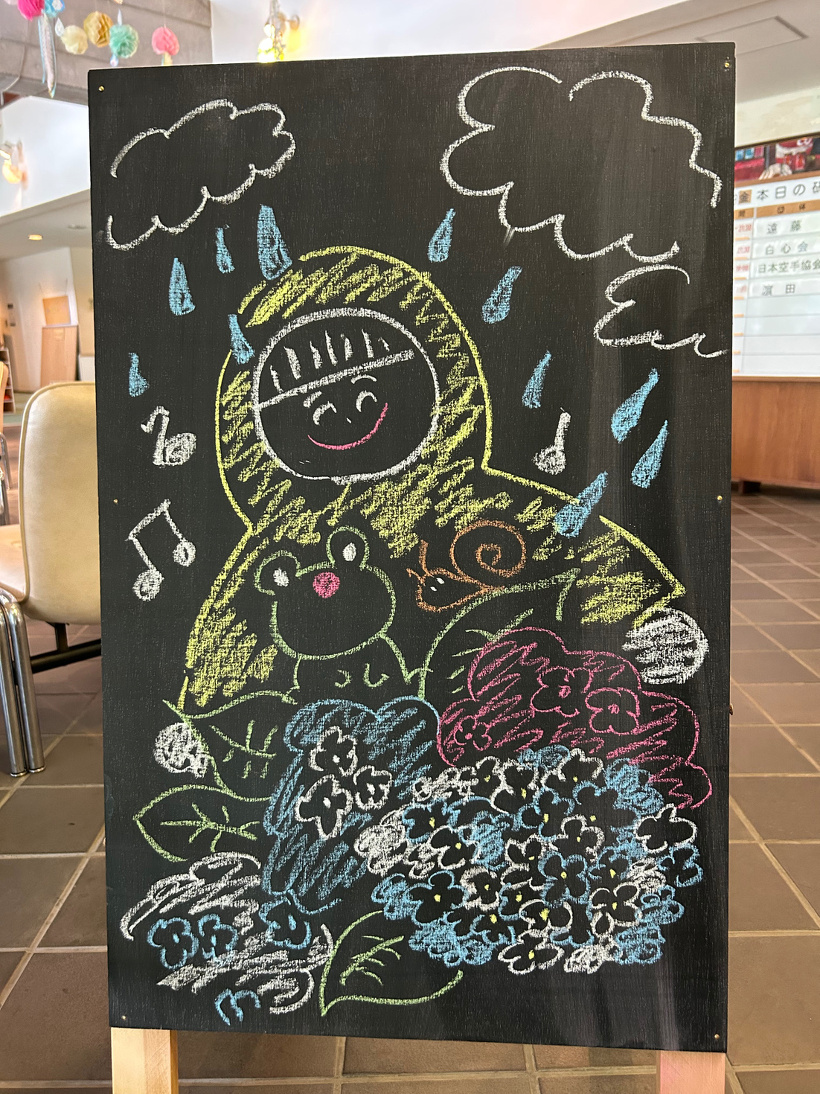 雨の中レインコートを着た笑顔の子ども、カエル、カタツムリ、アジサイをチョークで描いた黒板の立て看板
