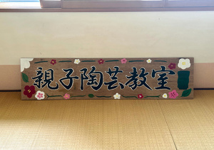 「親子陶芸教室」と書いた木の看板。白、赤、ピンクの花の絵で装飾されている。