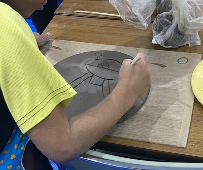 子どもが平らにした粘土に竹串のような道具でキャラクターを描いている様子